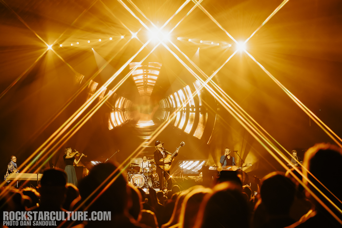 A live music performance with a band on stage, featuring musicians playing various instruments under bright, golden stage lights and visual projections in the background.