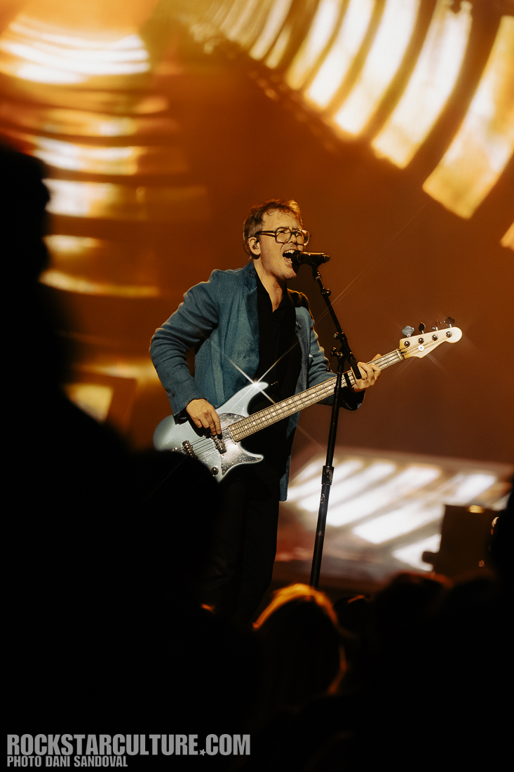 A musician performing on stage, singing into a microphone while playing a blue electric bass guitar, with a vibrant background of lights and patterns.