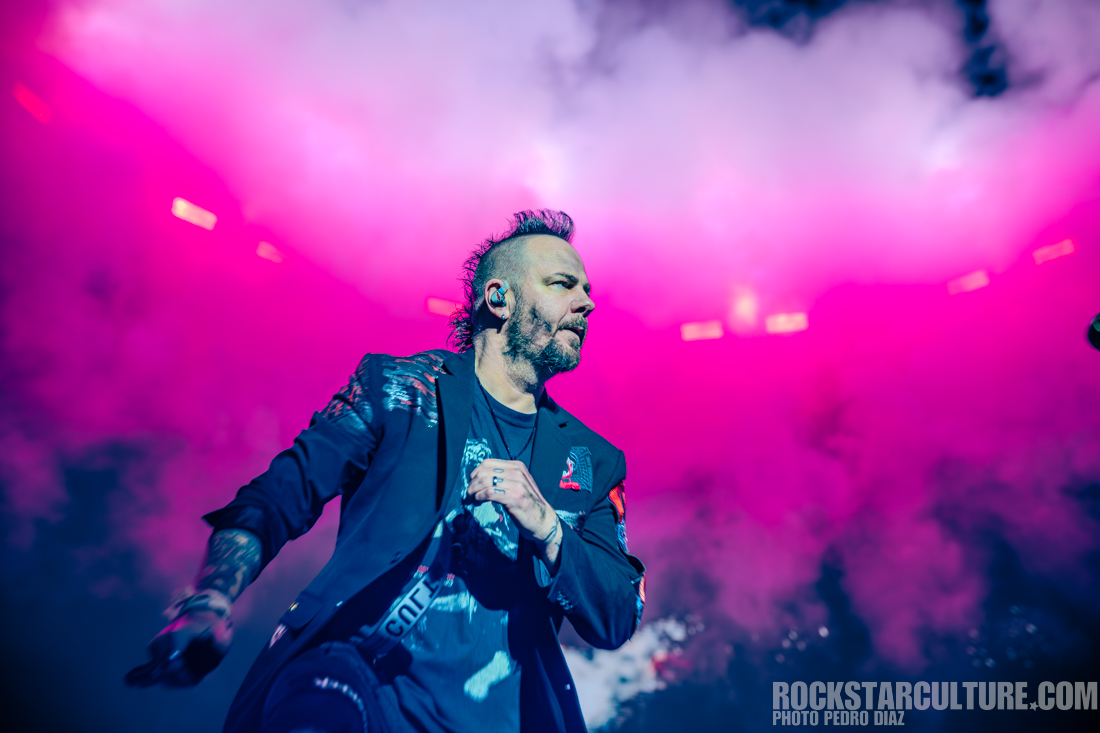 A male musician performs on stage surrounded by colorful smoke and vibrant lighting.