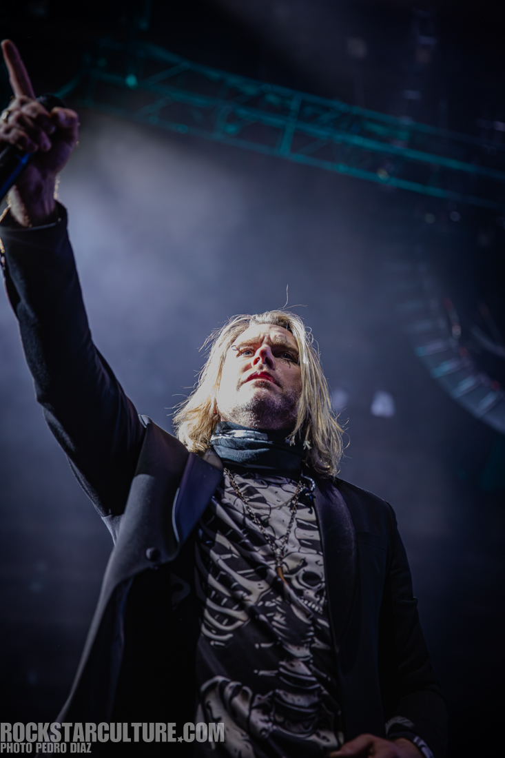 A male singer performing on stage with blonde hair and a black outfit, pointing towards the audience under dramatic lighting.