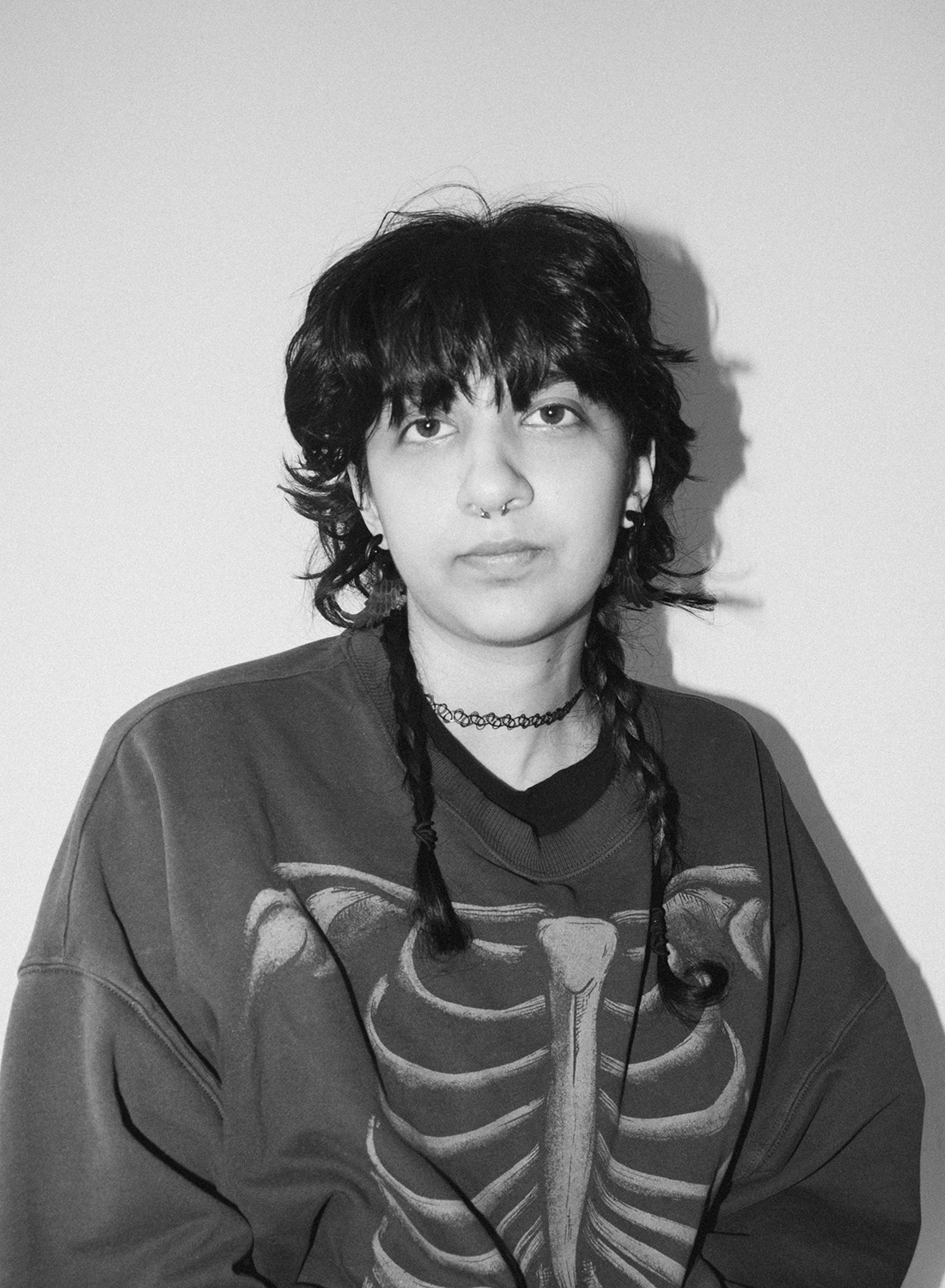 A person with dark, tousled hair styled in braids, wearing a black shirt with a skeleton graphic and a choker necklace, poses against a plain wall in black and white.