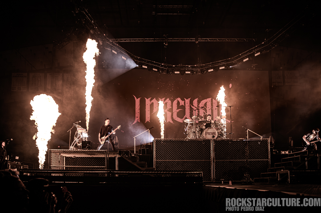 Live performance of a rock band on stage with amplified sound, featuring flames shooting up from the sides and a large backdrop displaying the band's name.