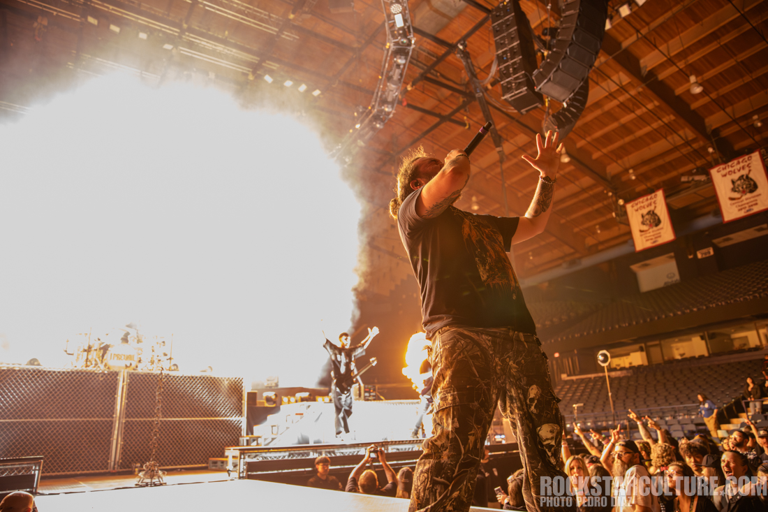 A male performer passionately singing on stage with a microphone, surrounded by flames and bright stage lights. In the background, band members play instruments, while the audience looks on excitedly.