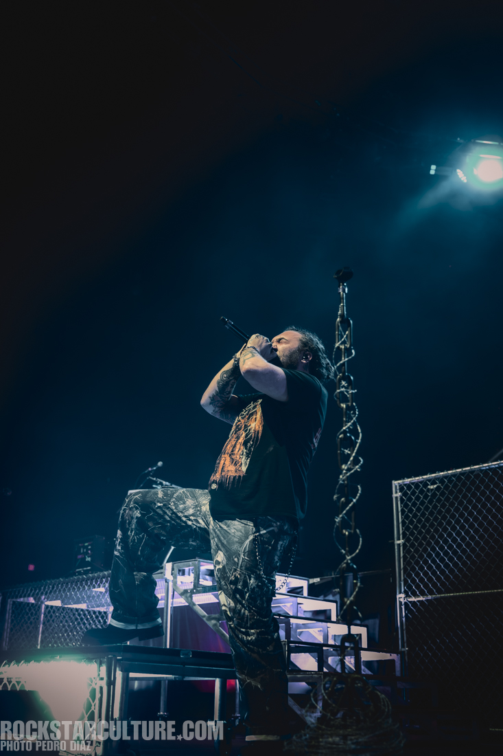 A musician performing on stage, passionately singing into a microphone with one foot elevated on a platform, dressed in a black shirt and camouflaged pants, with dramatic lighting in the background.