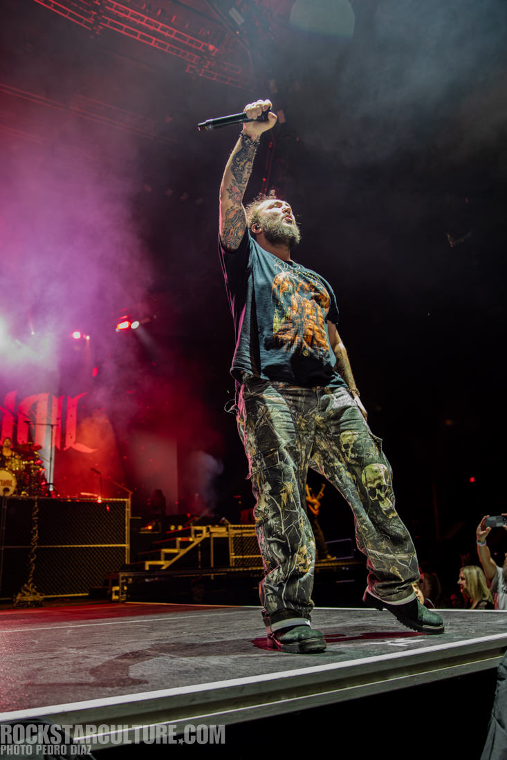 A performer on stage holding a microphone with one arm raised, dressed in a graphic t-shirt and camouflage pants, with smoke and colorful lights in the background.