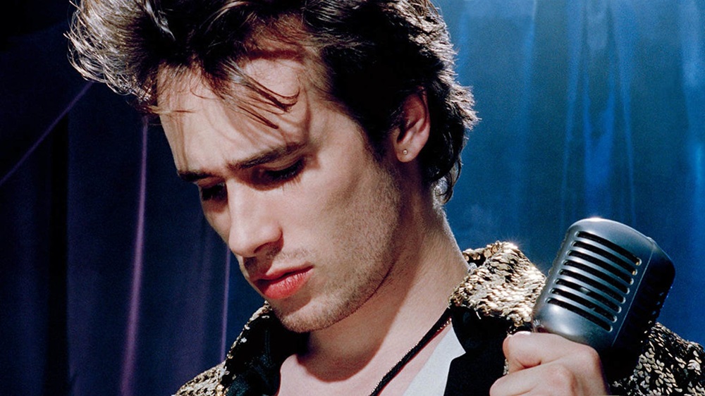 A young male singer with tousled hair, holding a vintage microphone, appears contemplative under soft blue lighting. He is wearing a stylish jacket and looks slightly downward.