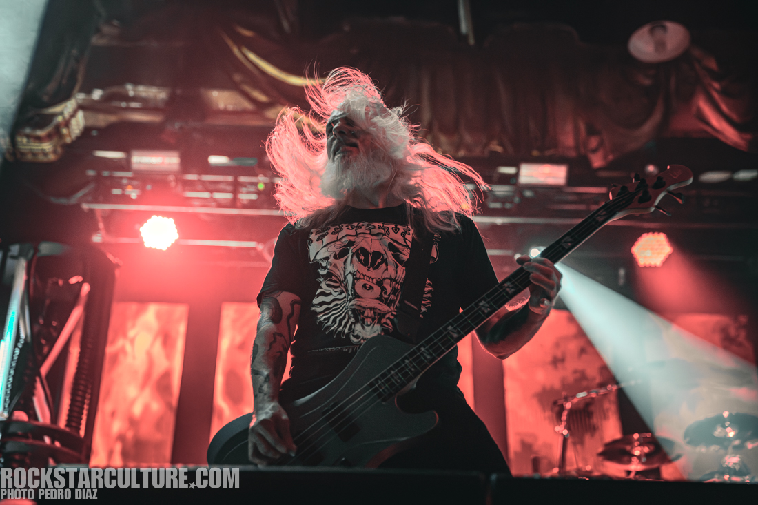 A musician with long hair and a beard plays a bass guitar on stage, illuminated by red and orange lights, wearing a graphic t-shirt featuring a skull and animal design.