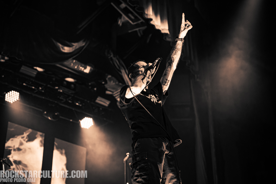 A singer performing on stage with one hand raised, holding a microphone, illuminated by stage lights in a concert setting.