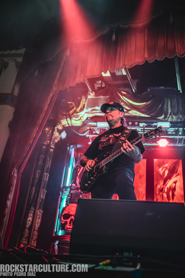 A guitarist performing on stage with dramatic lighting and a colorful backdrop featuring a skull design.