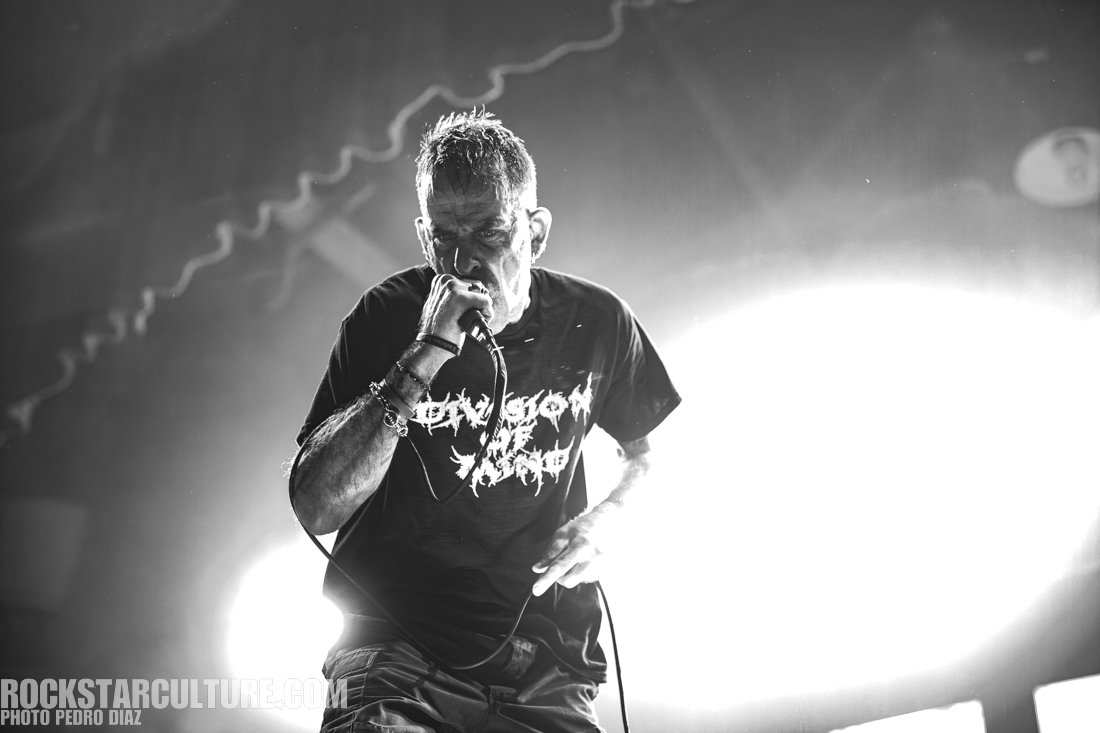 A vocalist passionately performing on stage, holding a microphone, wearing a black t-shirt with bold white lettering, set against a bright illuminated background.
