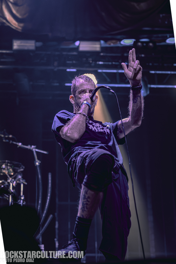 A male vocalist performs on stage, holding a microphone and gesturing with one hand while wearing casual black clothing.