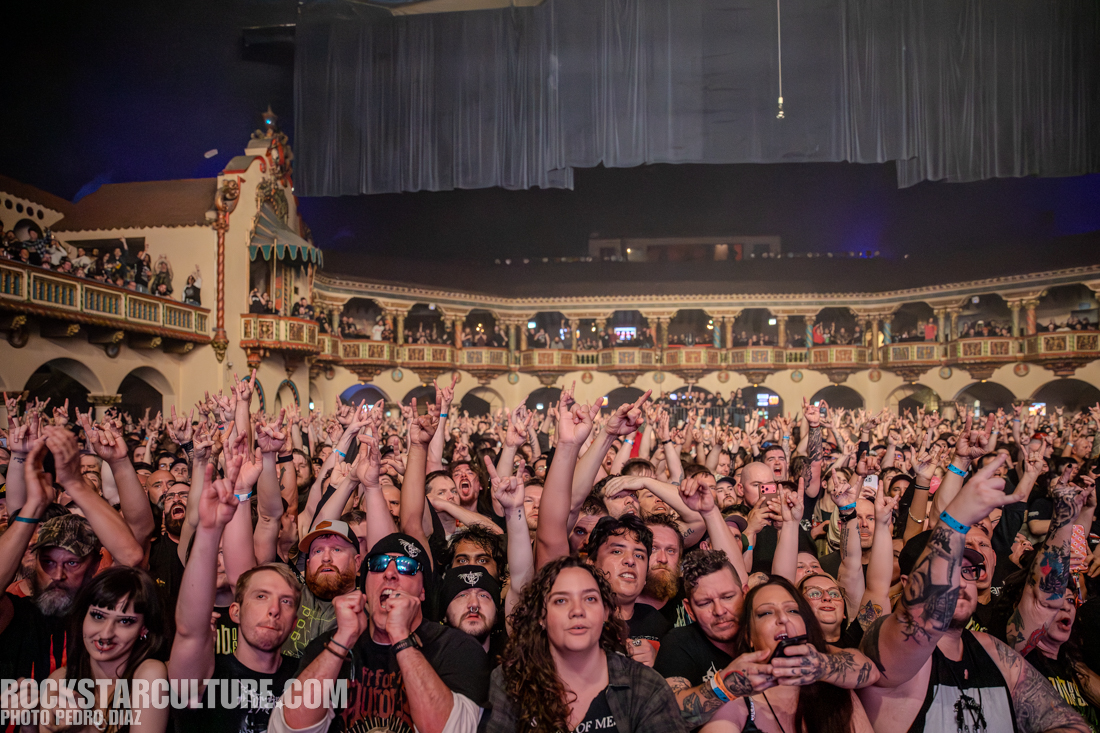 A lively concert crowd with raised hands and a variety of enthusiastic expressions, set in a historic venue with ornate decorations in the background.