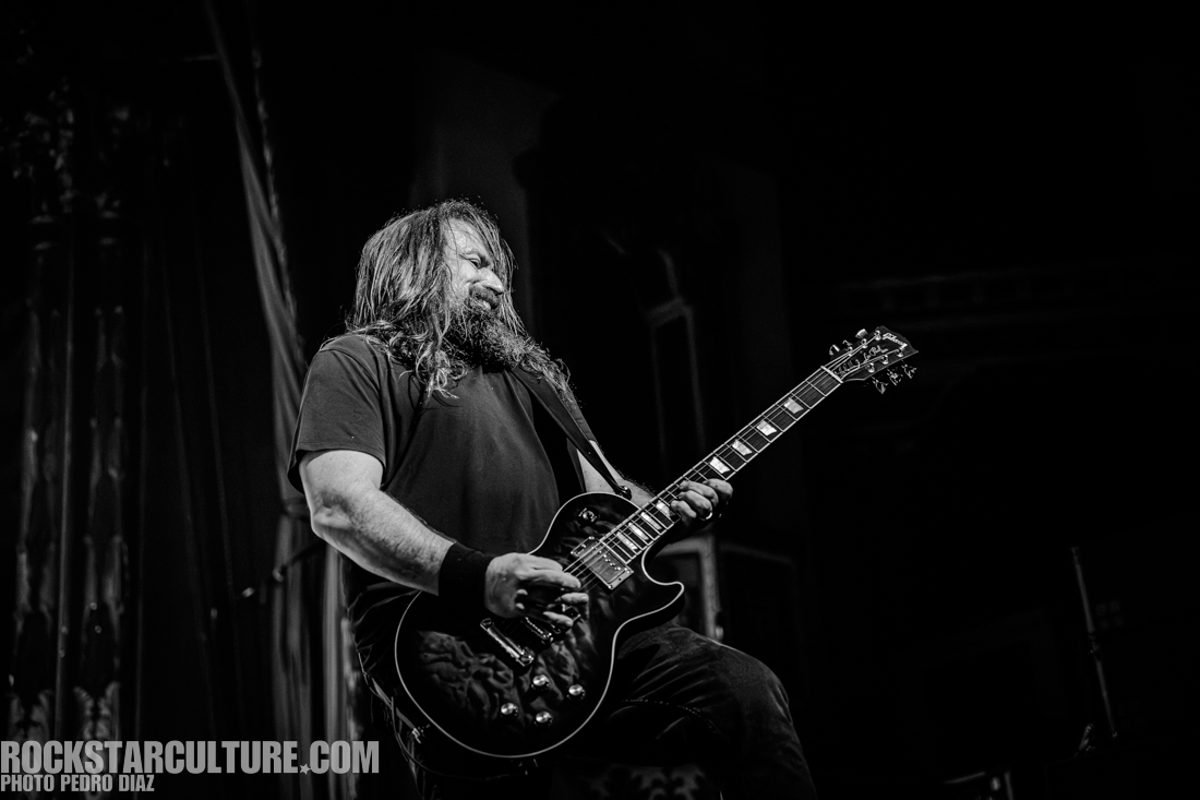A musician with long hair plays an electric guitar on stage, captured in black and white.