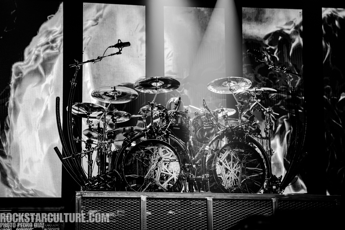 A black and white image of a drum set on stage, featuring multiple drums and cymbals, with an abstract background displayed on large screens.