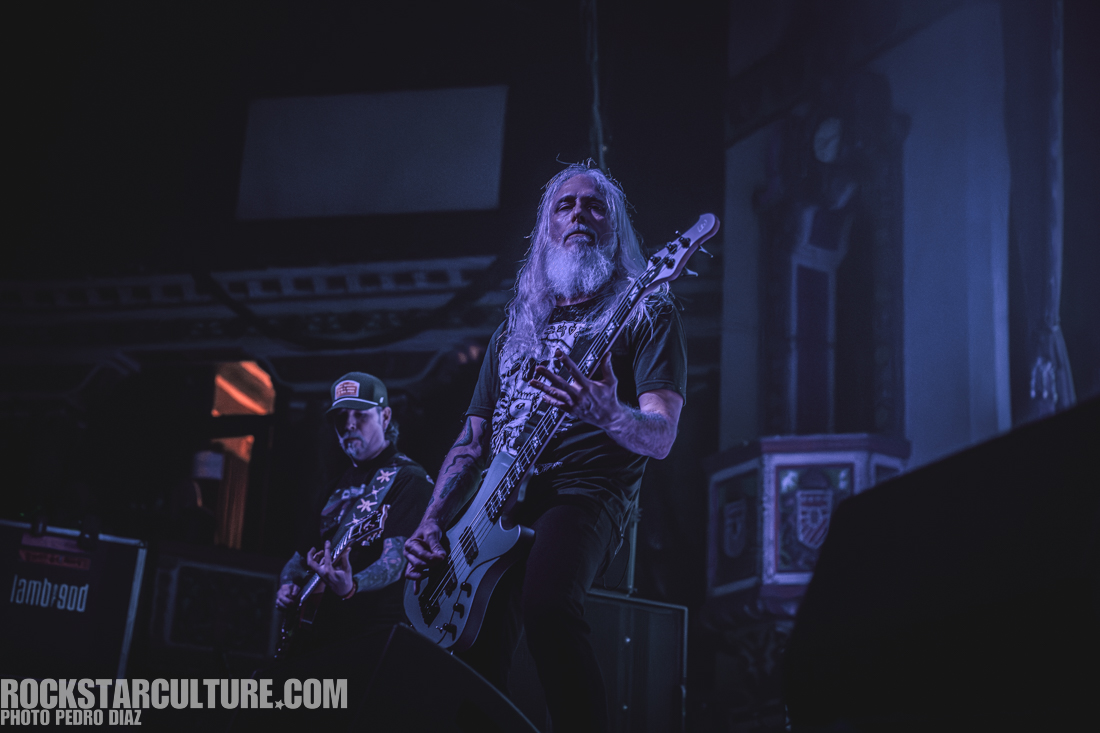 A musician with long gray hair and a beard plays a bass guitar on stage, while another band member in the background focuses on their instrument. The setting features dramatic lighting and a concert environment.