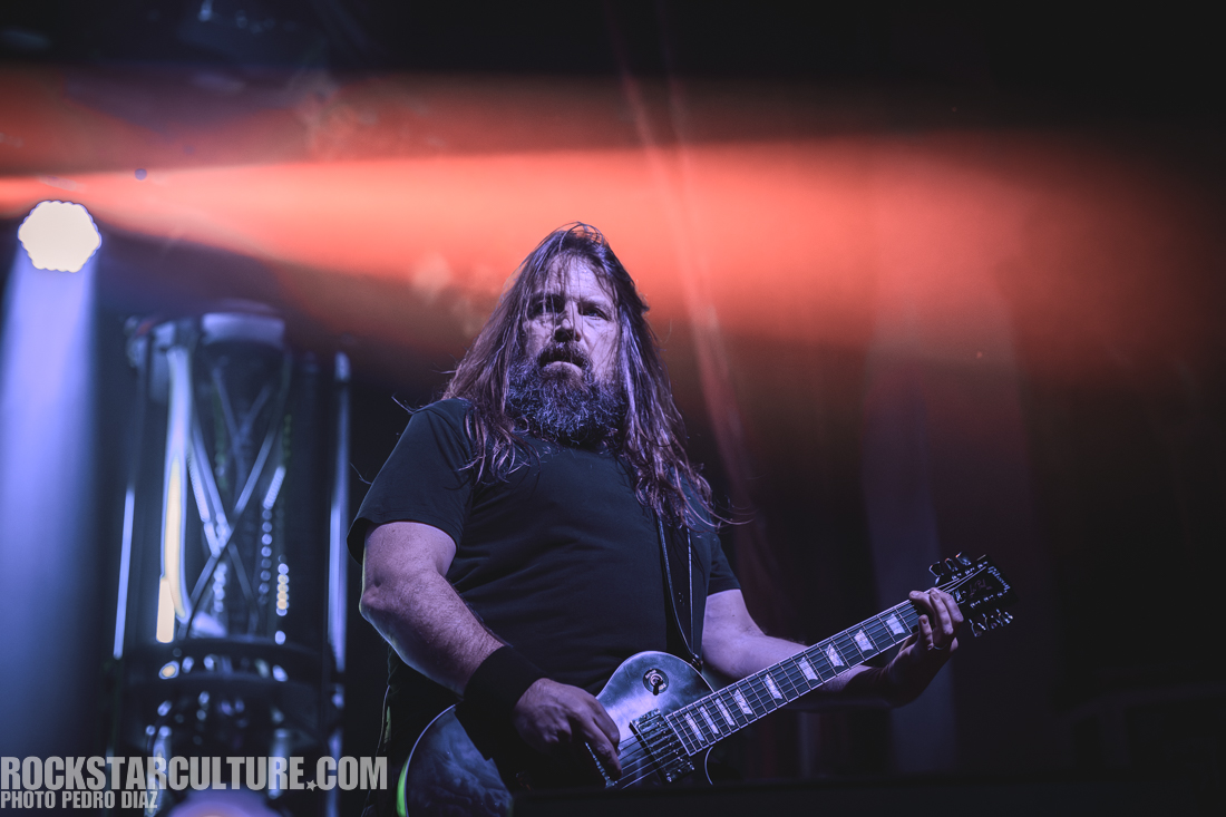 A guitarist performing on stage with a dramatic lighting backdrop, featuring long hair and a beard.