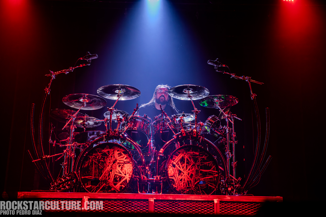 A drummer performs on stage surrounded by a vibrant display of red and blue lights, seated at an elaborate drum set.