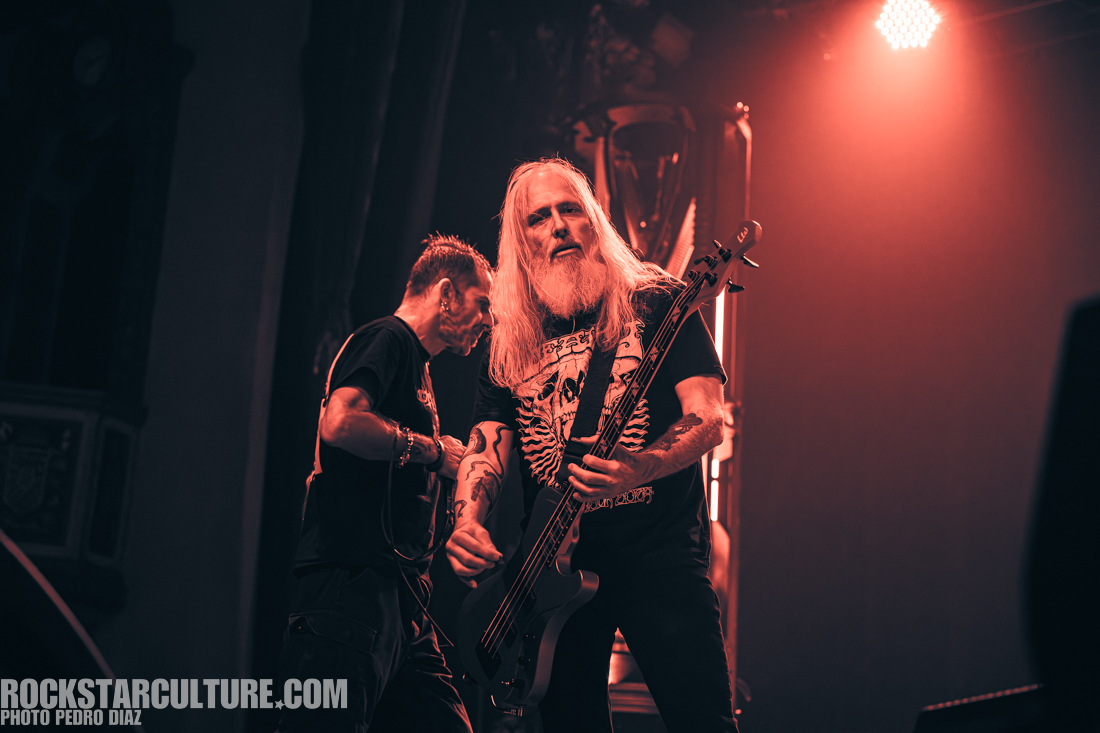 Two musicians performing on stage, one playing the bass guitar and the other standing nearby, in a vibrant, red-lit environment.