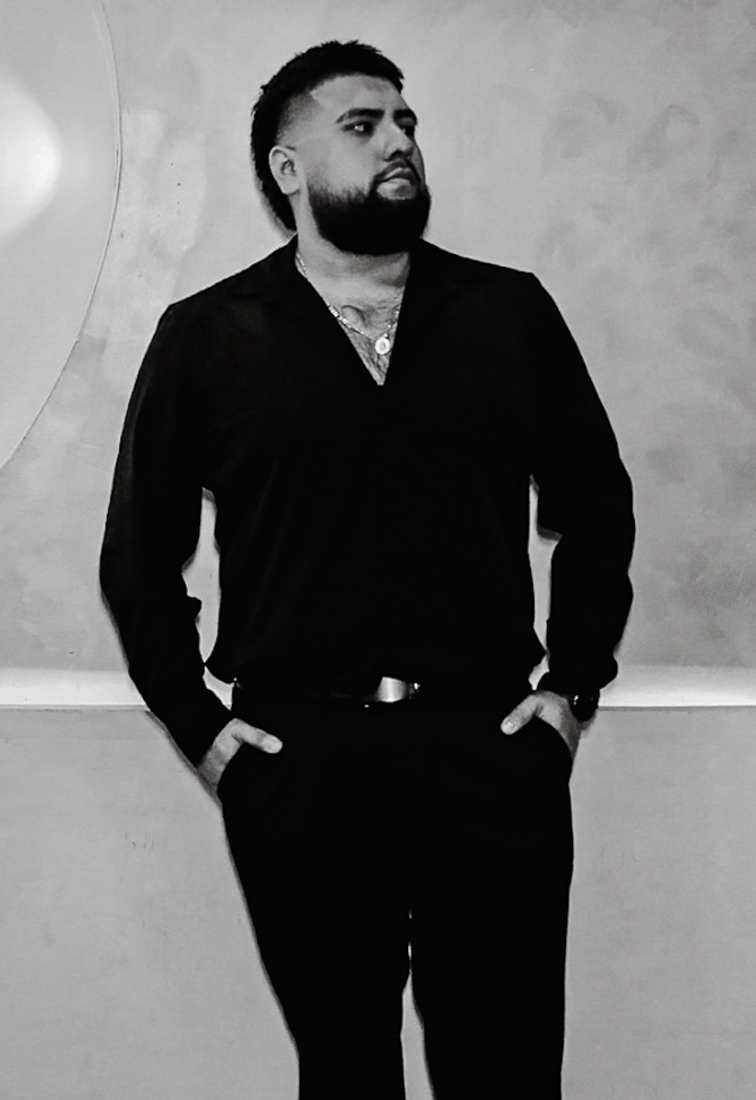 A man in a black button-up shirt and pants, standing confidently with his hands in his pockets against a neutral background.