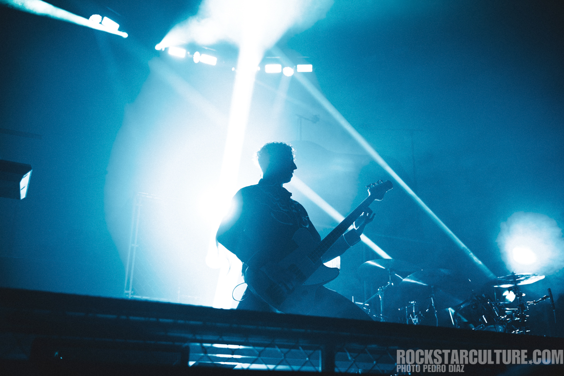 Silhouette of a guitarist performing on stage, illuminated by bright blue lights and smoke during a live concert.