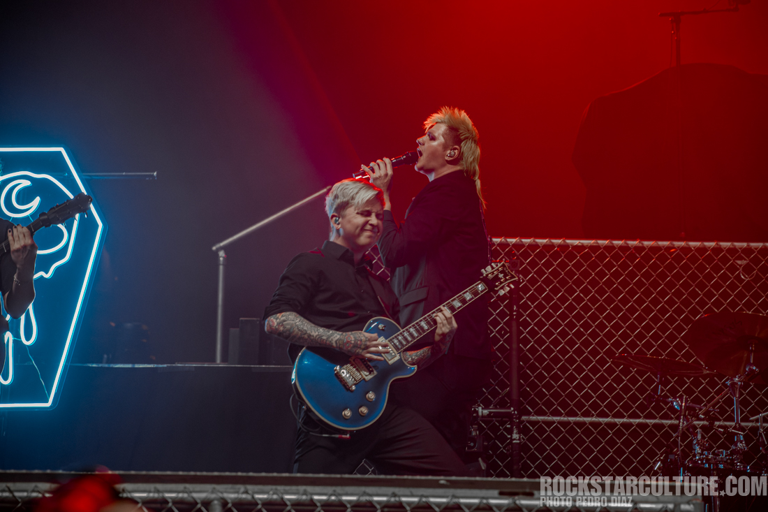 Two musicians performing on stage, one playing a blue electric guitar and the other singing into a microphone. A brightly lit geometric neon sign is visible in the background.