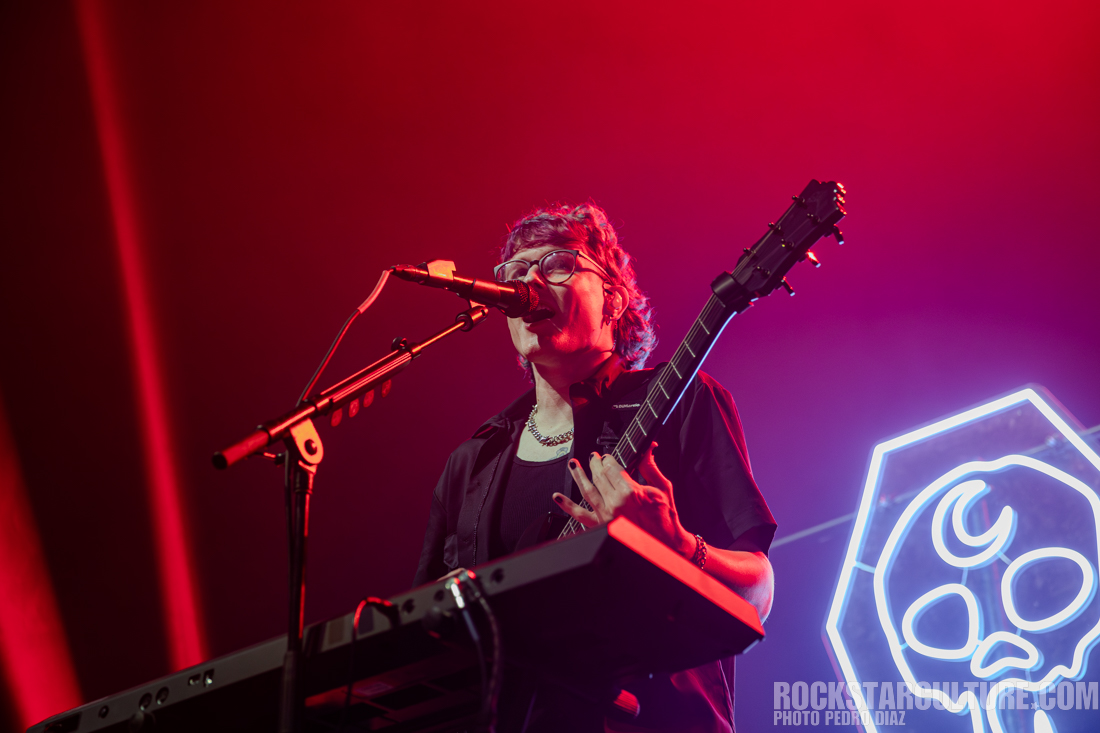 Musician performing on stage, holding a microphone and playing an electric guitar, with colorful stage lighting in the background.