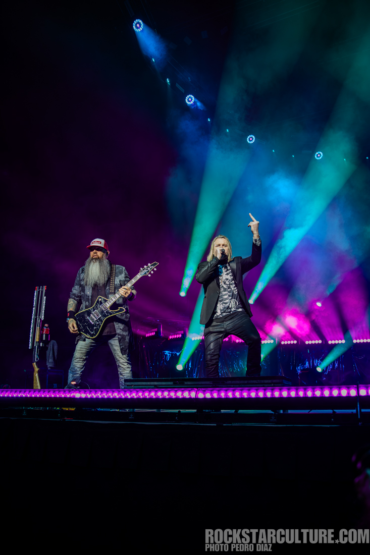 Two rock musicians perform on stage under colorful lights, one playing a guitar and the other singing passionately.