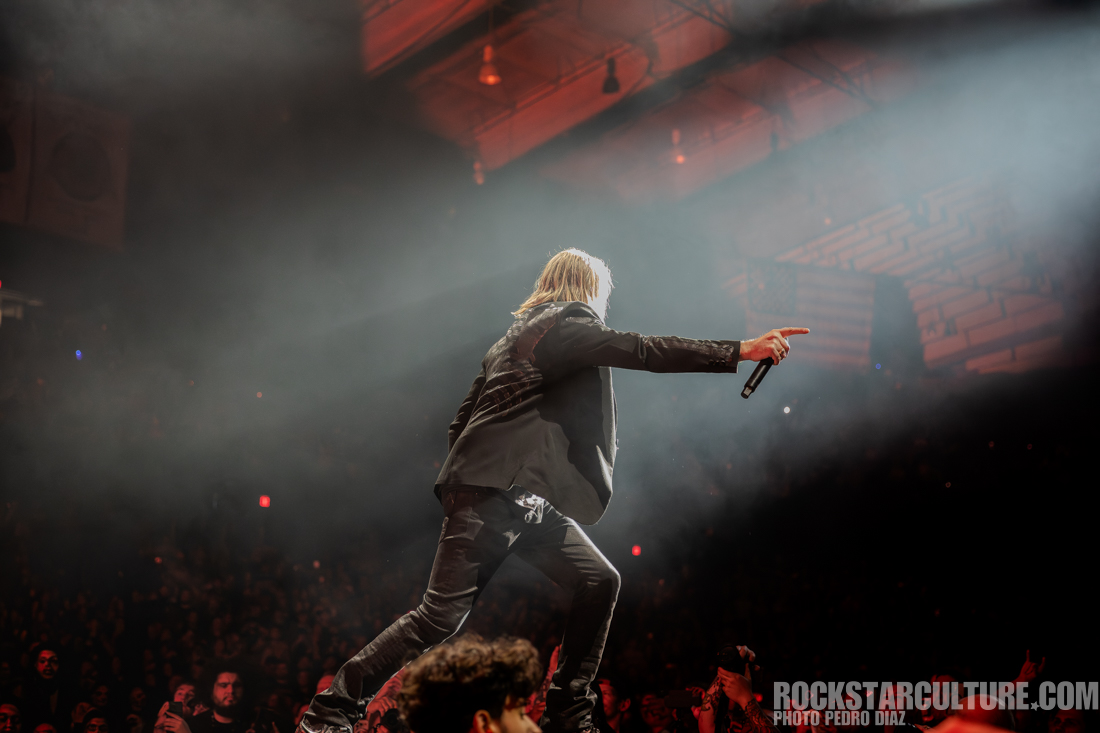 A performer on stage pointing out to the audience while holding a microphone, surrounded by dramatic lighting and fog.