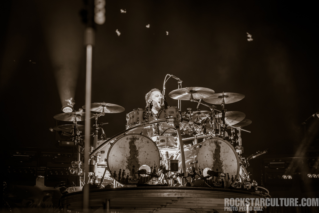A drummer performing on stage, surrounded by a set of drums with dramatic lighting and a dynamic atmosphere.