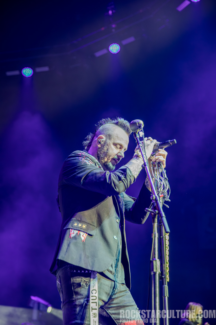A male singer performing on stage with a microphone, wearing a black jacket with embellishments, set against colorful stage lights.