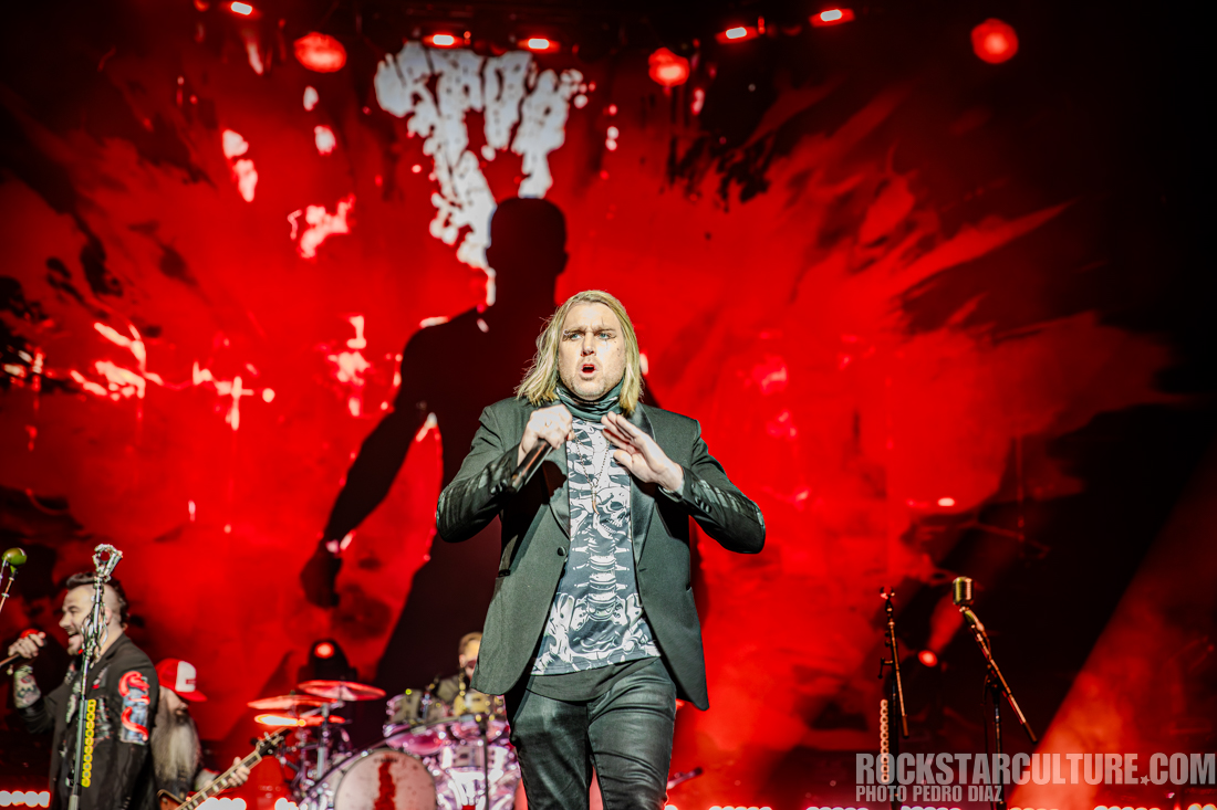 A male singer performing on stage with a dramatic red background and energetic stage lights.