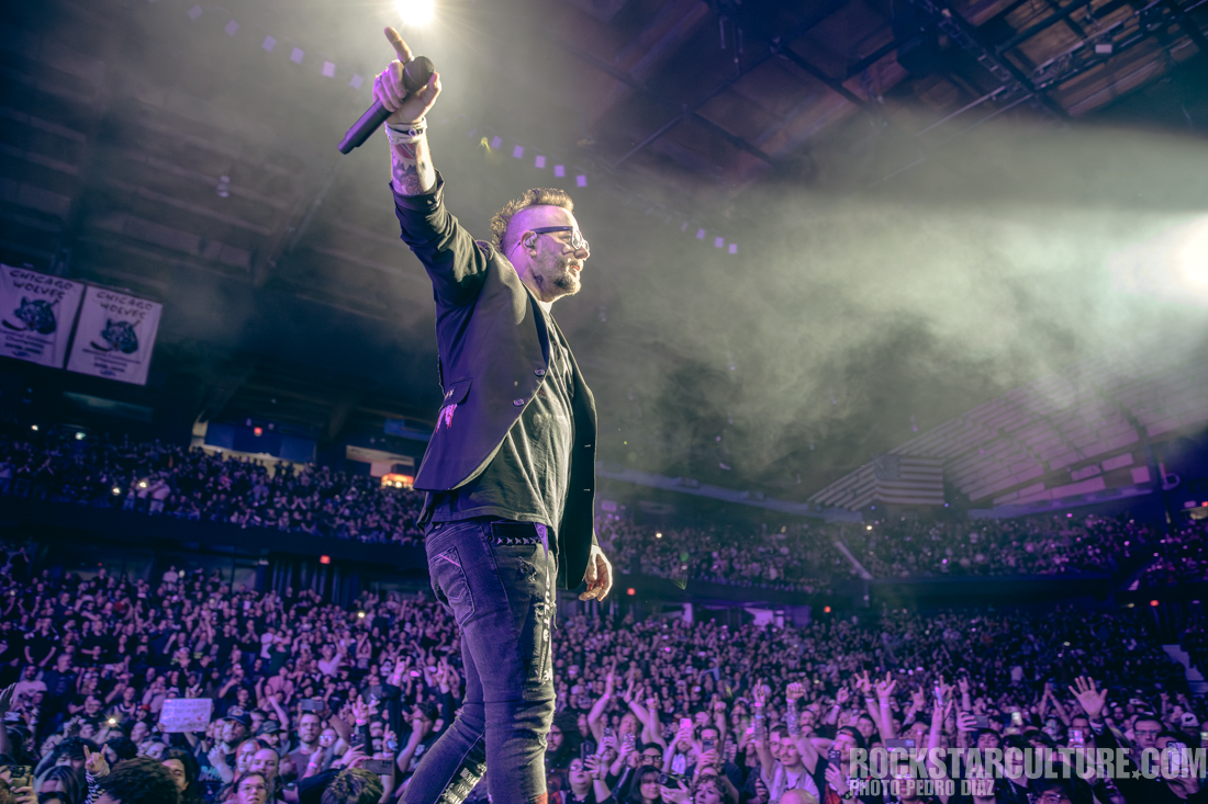 A male singer performing on stage with a microphone, gesturing to the audience in a packed arena, illuminated by stage lights and surrounded by a crowd of fans.