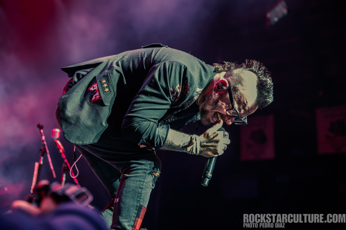 A performer in a black jacket with patches is passionately singing into a microphone on stage, with colorful lighting creating a dramatic atmosphere.
