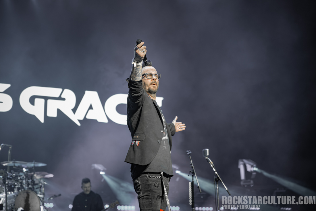 A musician performing on stage with arms raised, wearing a black outfit and glasses, with a large 'GRACE' sign in the background.