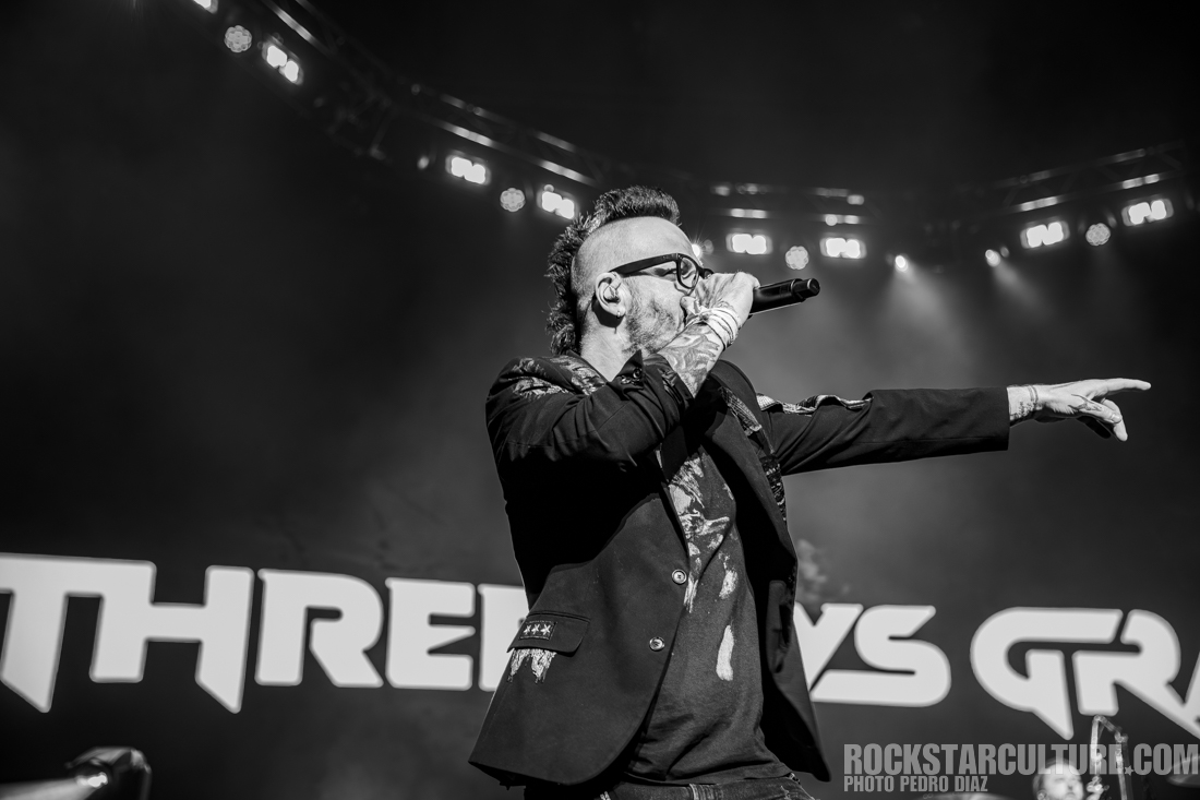 A male performer singing on stage, wearing a black jacket with designs, glasses, and tattoos visible on his arms, while directing the audience with his hand in a concert setting.