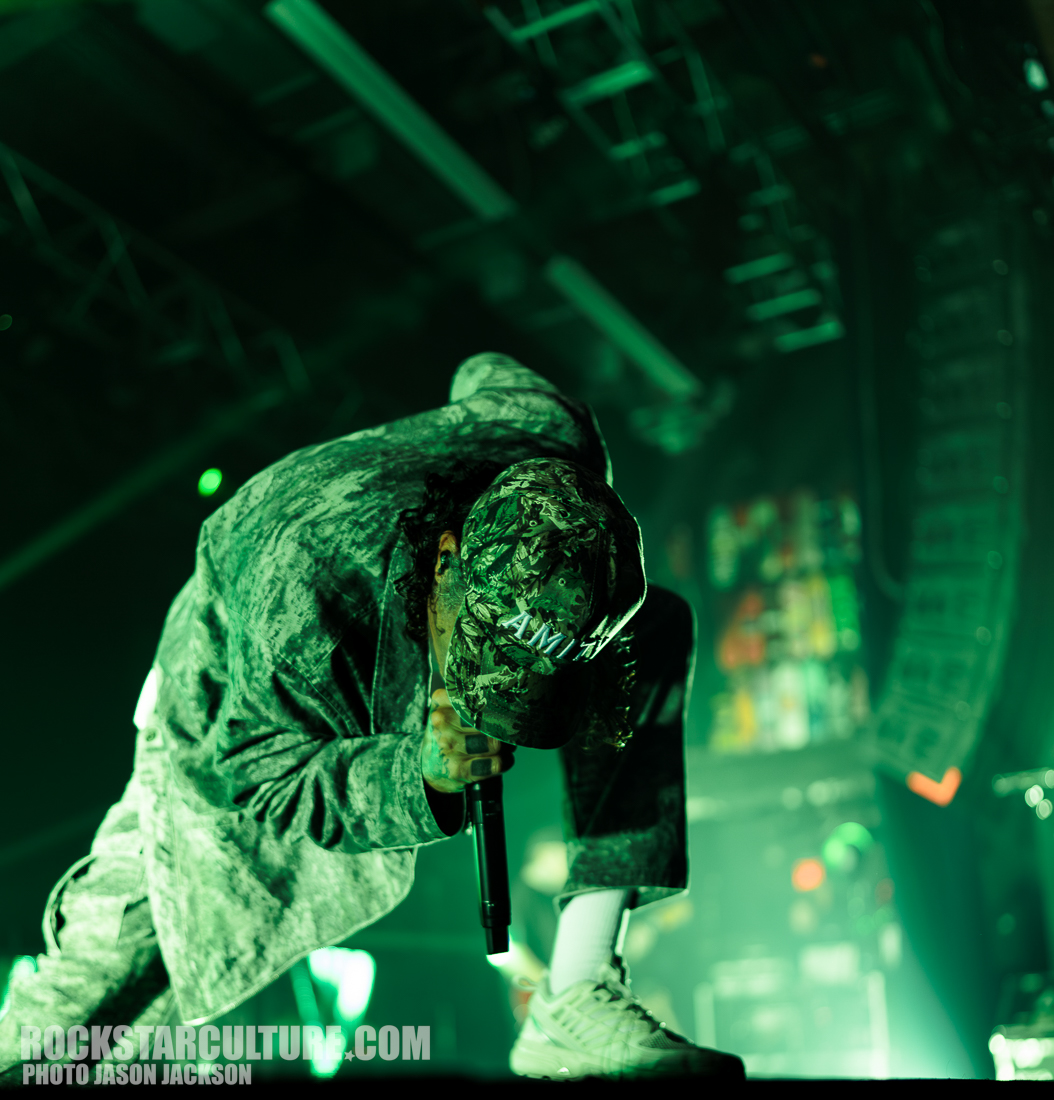 A performer crouches on stage, holding a microphone, with a focus on their energetic pose. They wear a camouflaged cap and a patterned outfit, under dramatic green stage lighting.