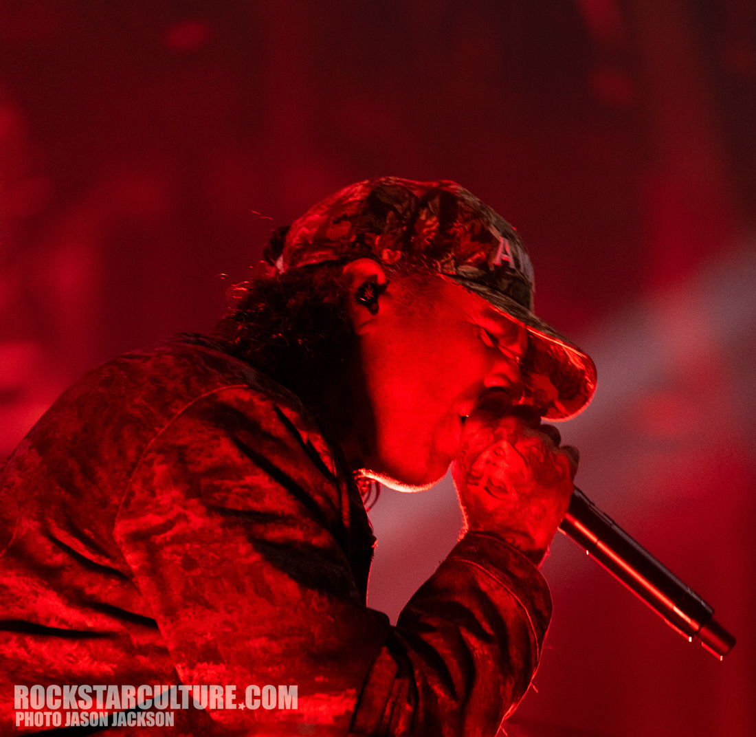 A performer singing into a microphone on stage, illuminated by red stage lights, wearing a camouflage cap and a dark jacket.
