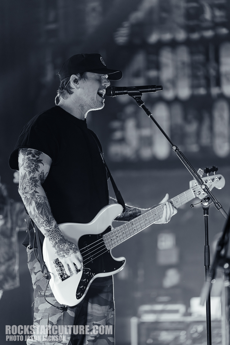 A musician performing on stage with a bass guitar, wearing a black t-shirt and a cap, with visible tattoos on his arms.