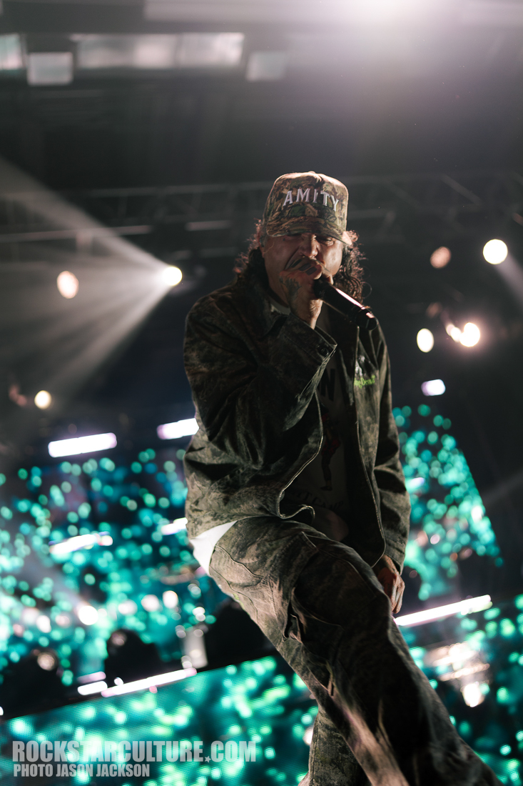 A performer wearing a camouflaged outfit and a cap, passionately singing into a microphone on stage, with colorful lights and a digital background illuminating the scene.