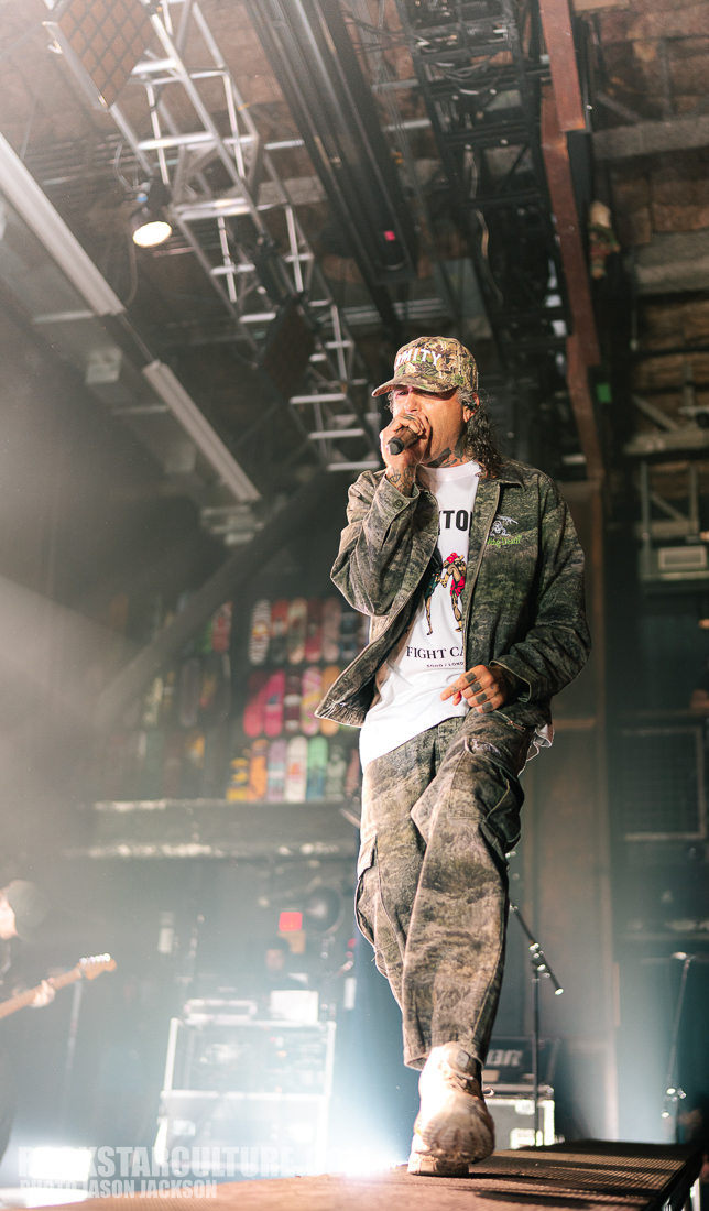 A performer on stage wearing a camouflage outfit and a hat, passionately singing into a microphone, with dramatic lighting and a blurred audience in the background.