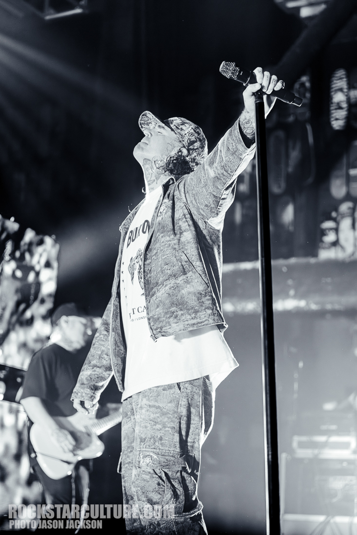 A male performer on stage wearing a patterned cap, gray jacket, and T-shirt, passionately singing into a microphone with one arm raised, while a guitarist is visible in the background.