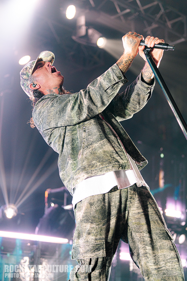 A musician passionately performing on stage, wearing a camouflage outfit and a cap, holding a microphone stand while singing.