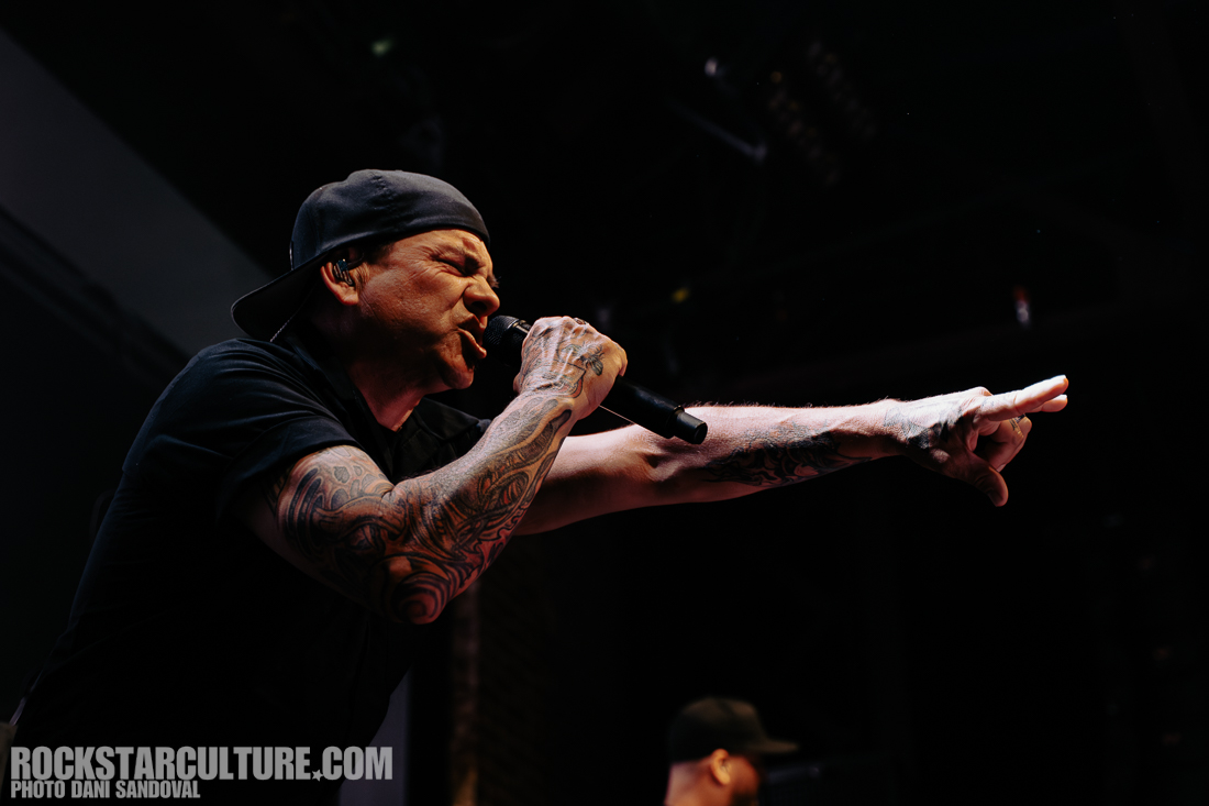 A male singer passionately performing on stage with a microphone, showcasing visible tattoos on his arms and wearing a black cap.