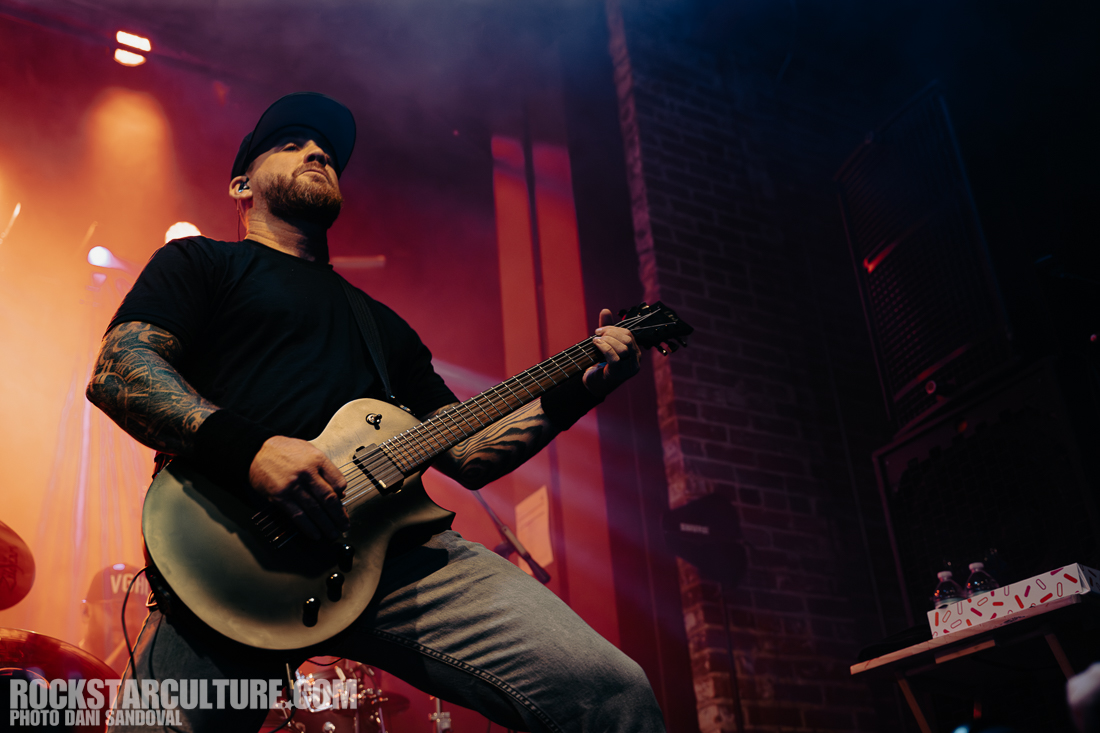 A guitarist performing on stage with a focused expression, wearing a black t-shirt and a cap, holding a light-colored guitar, surrounded by colorful stage lighting.