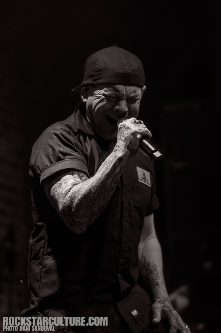 A male vocalist passionately performing on stage, wearing a cap and a dark shirt, with visible tattoos on his arms, in a dramatic black and white setting.