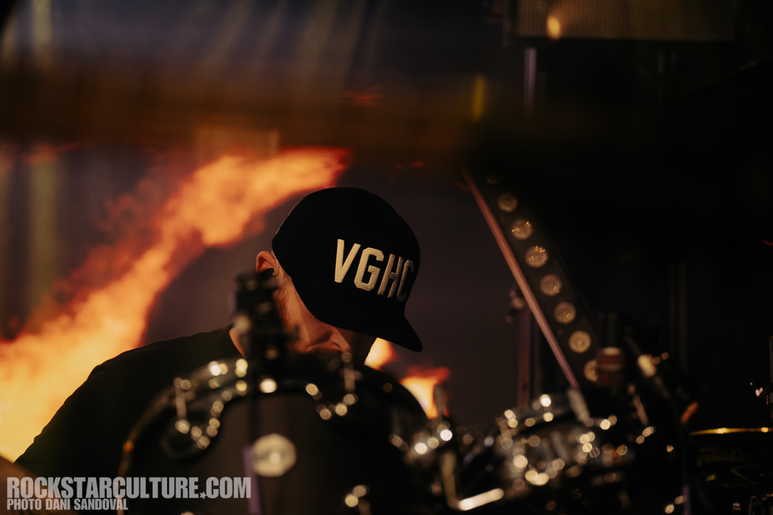 A musician wearing a black cap with 'VGHC' embroidered on it, focused on their instrument in a performance setting, with vibrant lights and a blurred background.