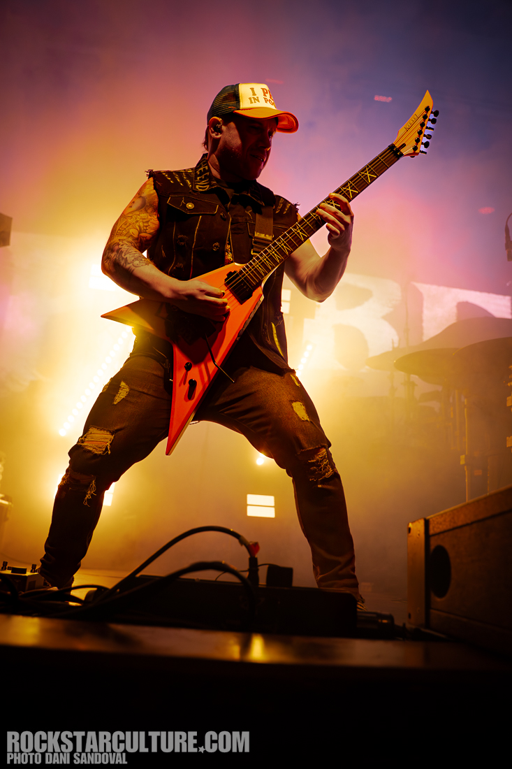 A guitarist performing on stage, dressed in a sleeveless jacket and a cap, playing an orange electric guitar with colorful stage lighting in the background.