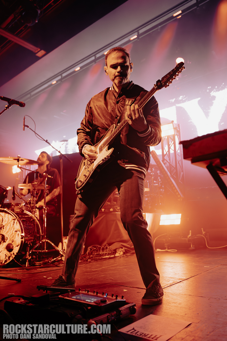 A musician playing an electric guitar on stage during a live performance, with warm stage lighting and a drummer visible in the background.