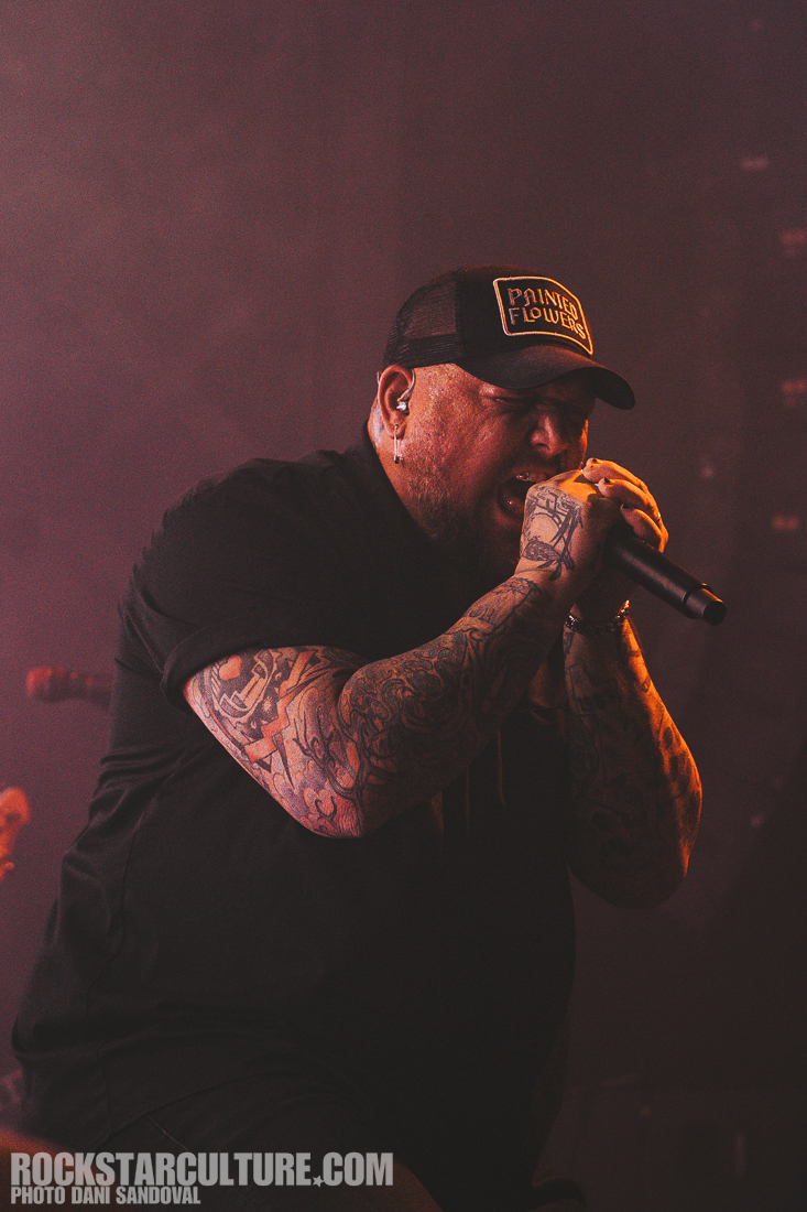 A vocalist passionately performing on stage, wearing a black shirt and cap, showcasing multiple tattoos on their arms, with a microphone in hand and dramatic lighting in the background.