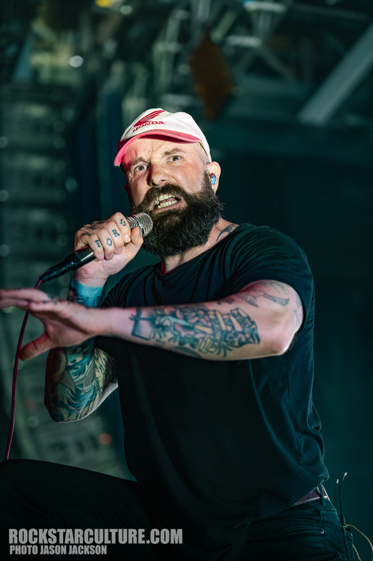 A vocalist on stage passionately performing into a microphone, wearing a white cap and a black t-shirt, showcasing various tattoos on his arms.